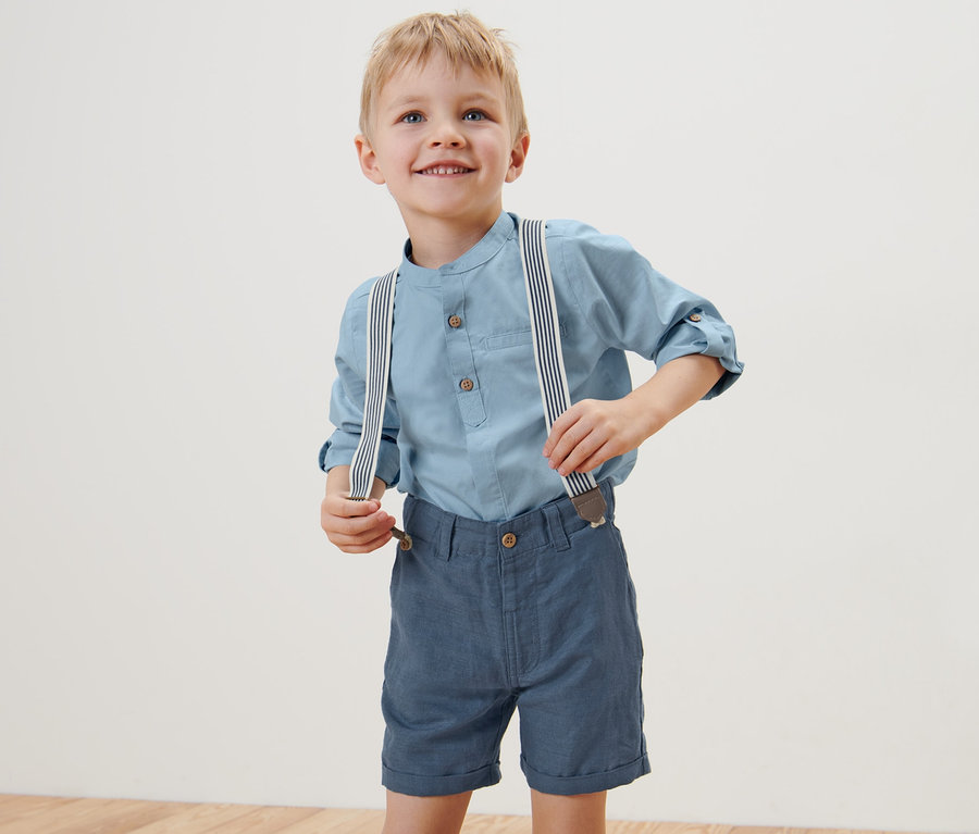 Ein Junge trägt ein blaues Hemd und blaue Kinder-Shorts und Hosenträger und lächelt.