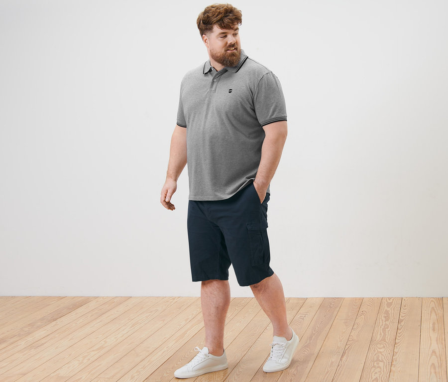 Ein Mann geht auf einem Holzboden und trägt ein graues Piqué-Poloshirt und navy Cargo-Shorts.