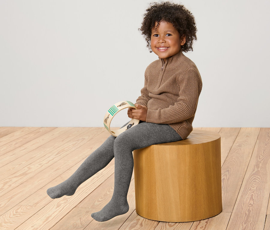 Ein lächelndes Kind sitzt auf einem Holzhocker und hält ein Tamburin. Es trägt einen braunen Pullover und eine graumelierte 2 Kinder-Thermostrumpfhosen, meliert.