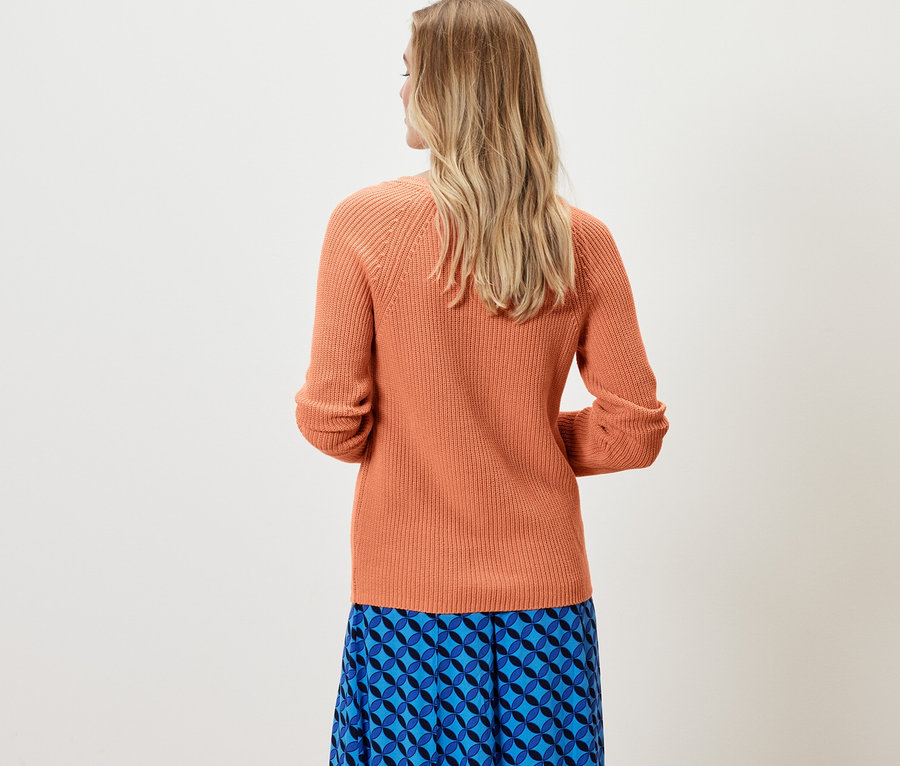 Frau steht mit dem Rücken zur Kamera und trägt einen peachfarbenen Strickpullover und einen blauen Rock.