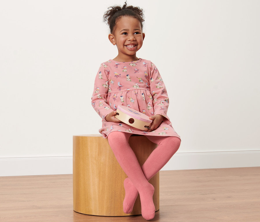 Lächelndes kleines Mädchen sitzt auf einem Holzhocker und hält ein Tamburin. Sie trägt ein rosa Kleid mit Print und 2 Kleinkind-Thermostrumpfhosen, Vogel.