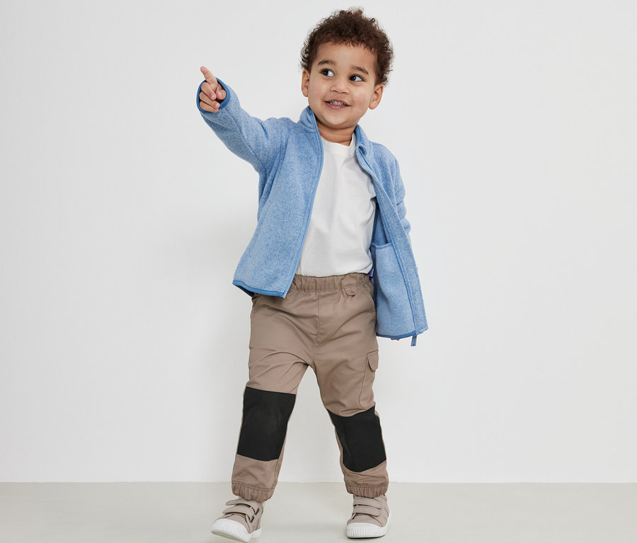 Junge zeigt mit dem Finger, trägt eine Kinder-Outdoorhose, eine blaue Fleecejacke und ein weißes T-Shirt.