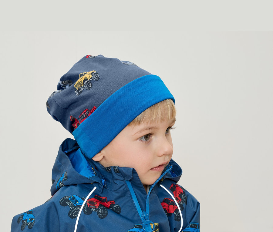 Ein Junge mit blonden Haaren trägt eine blaue Mütze und eine Jacke mit Monster-Truck-Aufdruck.