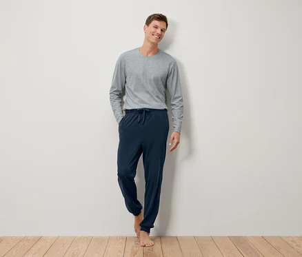 Ein barfüßiger Mann steht an einer Wand in einem grauen Langarmshirt und einer marineblauen Jogginghose.