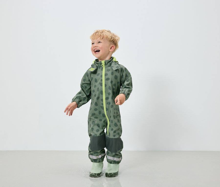 Ein Junge im grünen Overall mit Fleckenmuster und grünen Gummistiefeln springt in eine Pfütze.