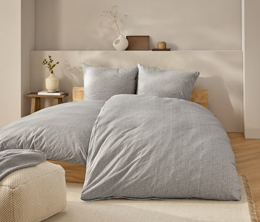 Bett mit grauer Bettwäsche und zwei Kissen. Auf dem Bett liegt eine graue Decke. Neben dem Bett steht ein beiger Pouf.