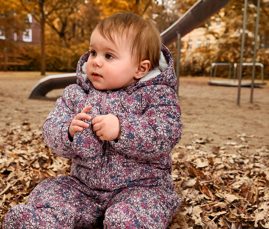 Baby sitzt in Baby-Winteroverall auf Blättern auf einem Spielplatz.