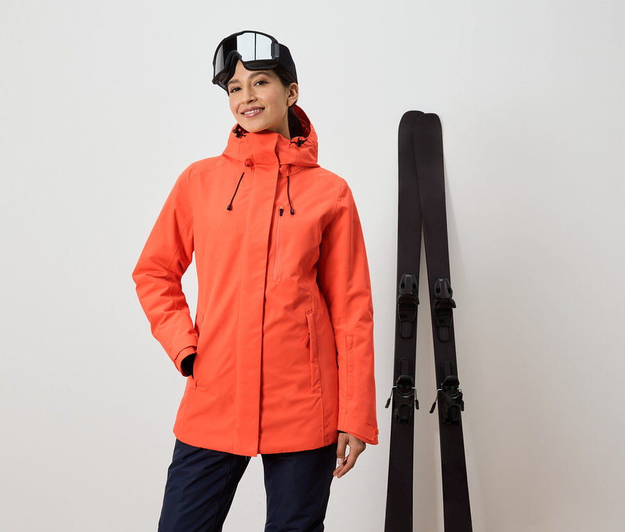 Frau mit orangefarbener Jacke und Skibrille steht neben Skiern.