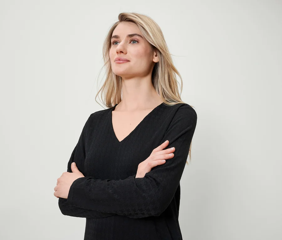 Blonde Frau mit verschränkten Armen trägt ein schwarzes Langarmshirt mit V-Ausschnitt.
