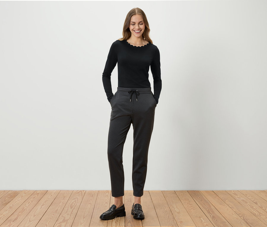 Eine Frau steht und lächelt, sie trägt ein schwarzes Langarmshirt, eine graue gemusterte Hose und schwarze Slipper.