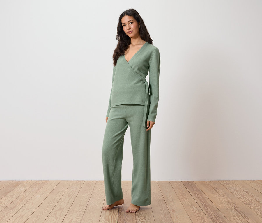 Frau in grünem Wickeloberteil und Hose steht auf Holzboden.