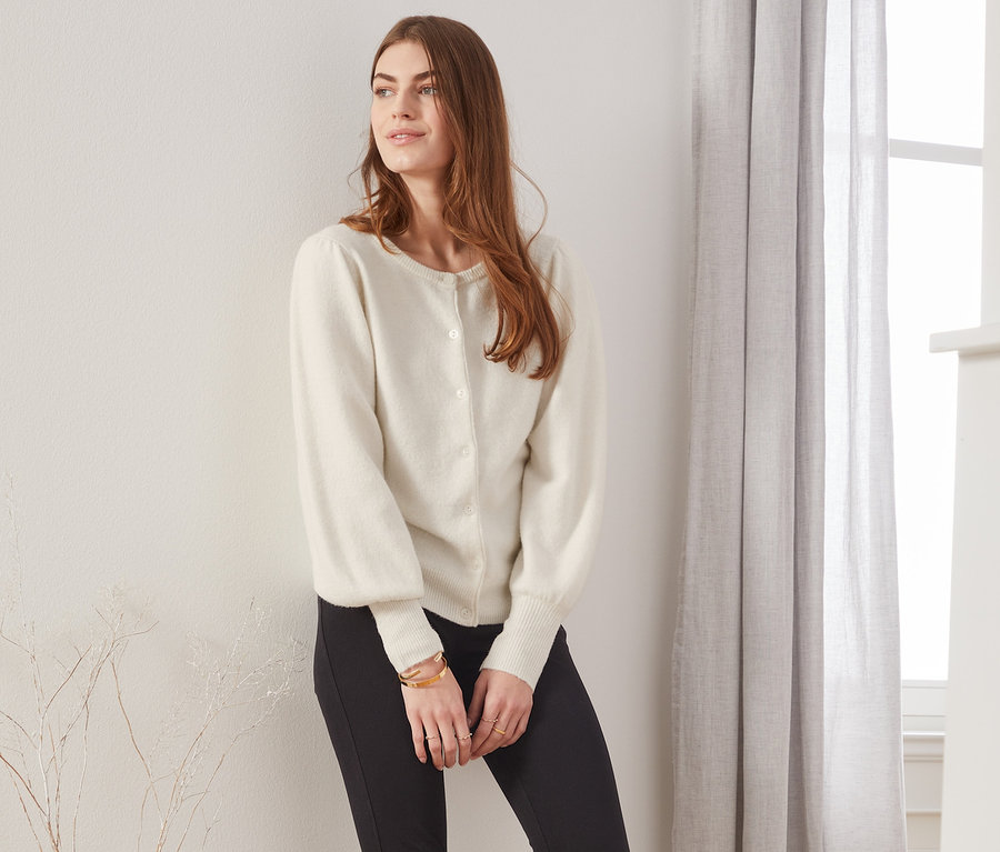 Eine Frau, die an einer Wand lehnt, trägt eine cremefarbene Feinstrickjacke mit Puffärmeln, eine schwarze Hose und Stiefel.