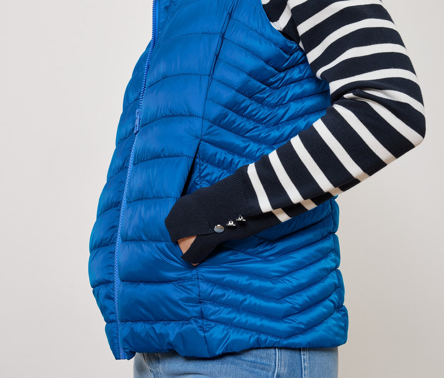 Person mit Hand in der Tasche einer blauen Steppweste und einem marineblau-weiß gestreiften Hemd.