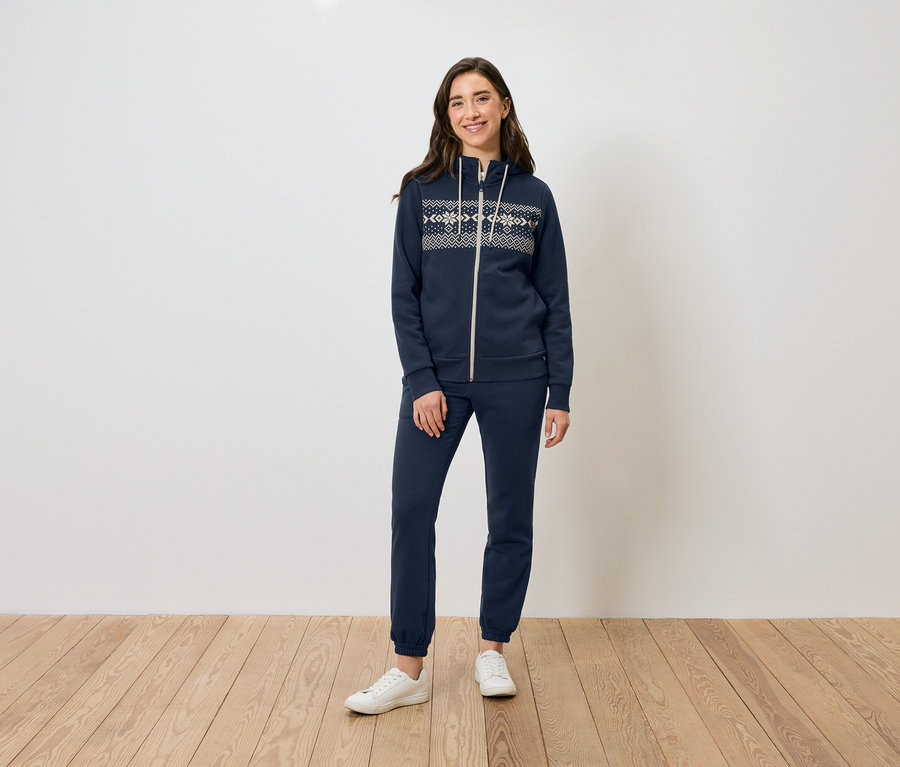 Eine Frau steht auf einem Holzboden und trägt einen dunkelblauen Reißverschluss-Hoodie und eine Trainingshose sowie weiße Turnschuhe.