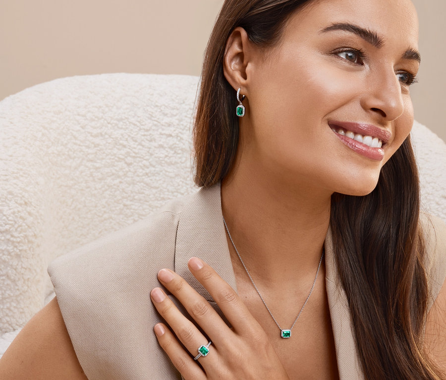 Lächelnde Frau sitzt im Sessel und trägt Ohrringe, Halskette und Ring mit smaragdgrünen Steinen.