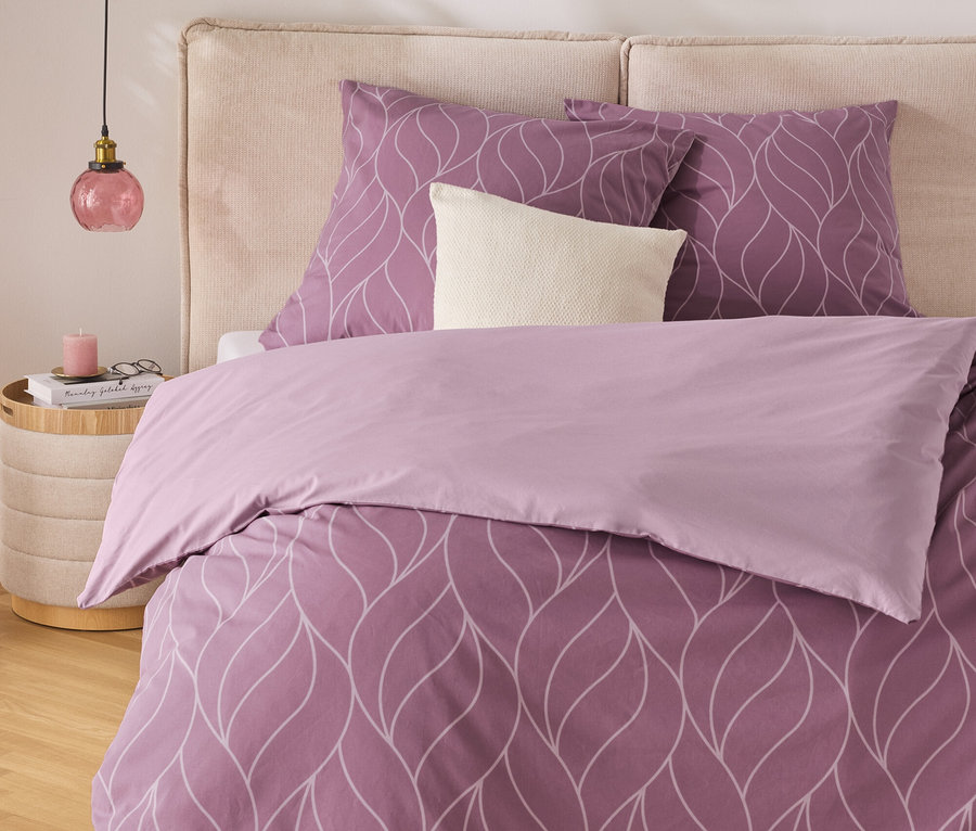 Lila Bettwäsche mit weißem Blattmuster auf einem Bett mit beigem Kopfteil. Ein Nachttisch mit Lampe steht neben dem Bett.