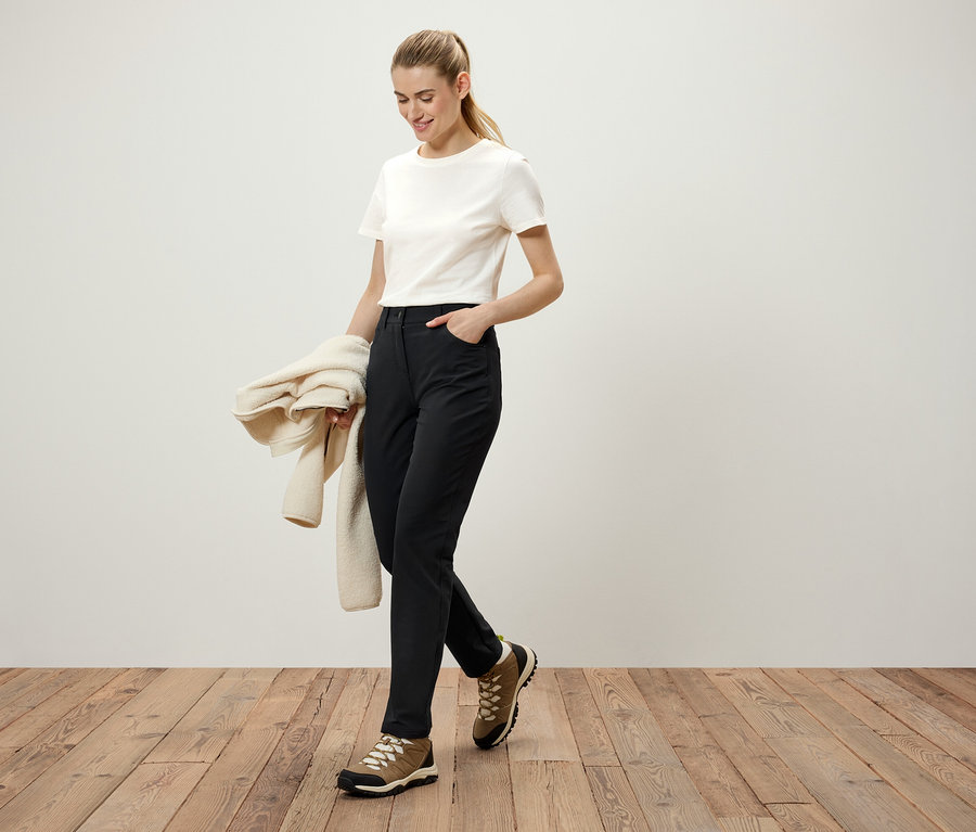 Frau geht auf einem Holzboden, trägt eine schwarze Hose, ein weißes T-Shirt und braune Schuhe. Sie hält einen cremefarbenen Pullover in der Hand.