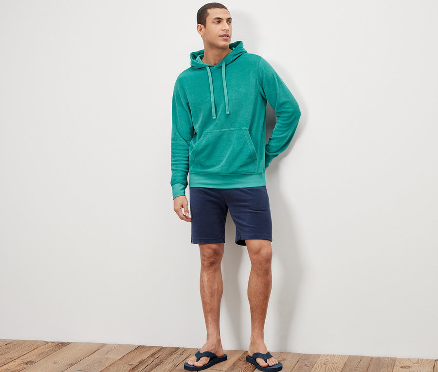 Mann steht und trägt einen Frottee-Hoodie, Shorts und Flip-Flops.