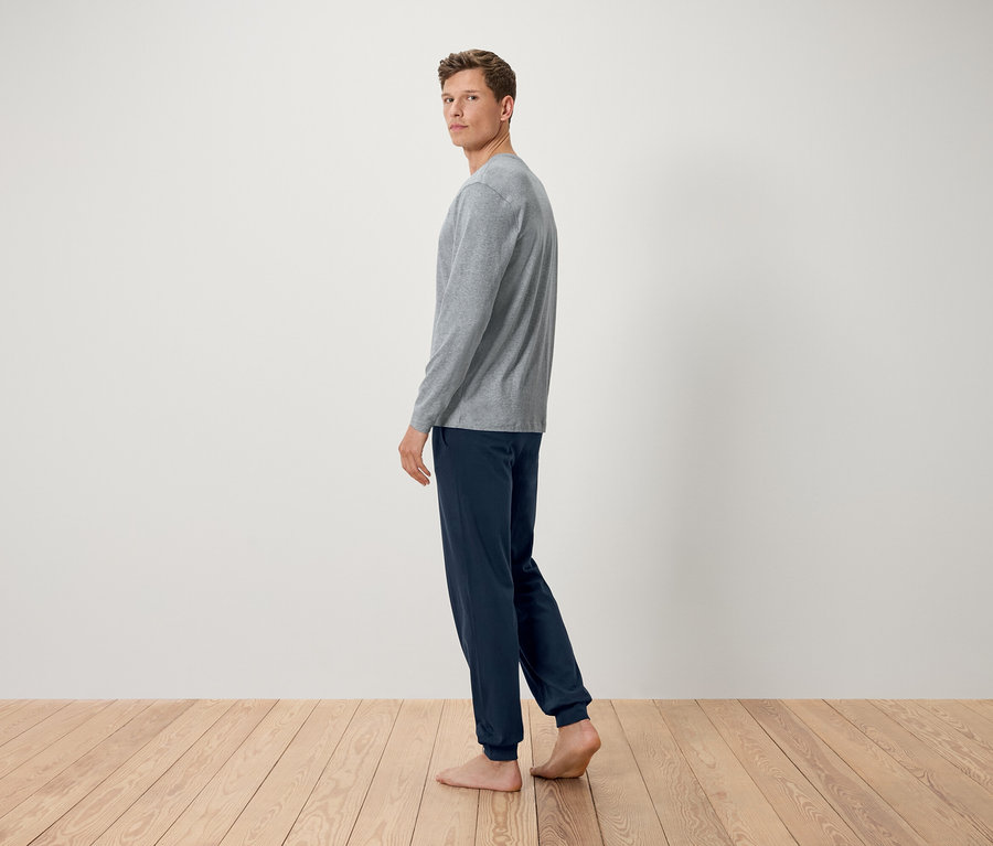 Ein Mann in einem grauen Langarmshirt und dunkelblauen Hosen steht barfuß auf einem Holzboden und blickt über seine Schulter.