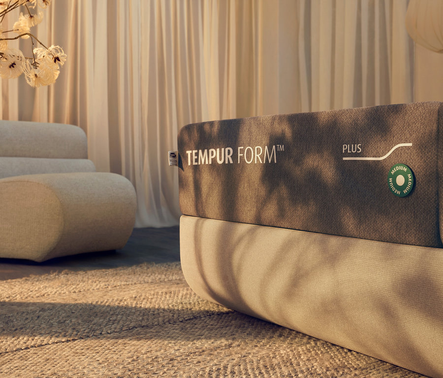 Nahaufnahme einer TEMPUR Form™ Plus Hybridmatratze auf einem Bett mit beigem Kopfteil. Im Hintergrund befinden sich ein beiger Sessel und Vorhänge.