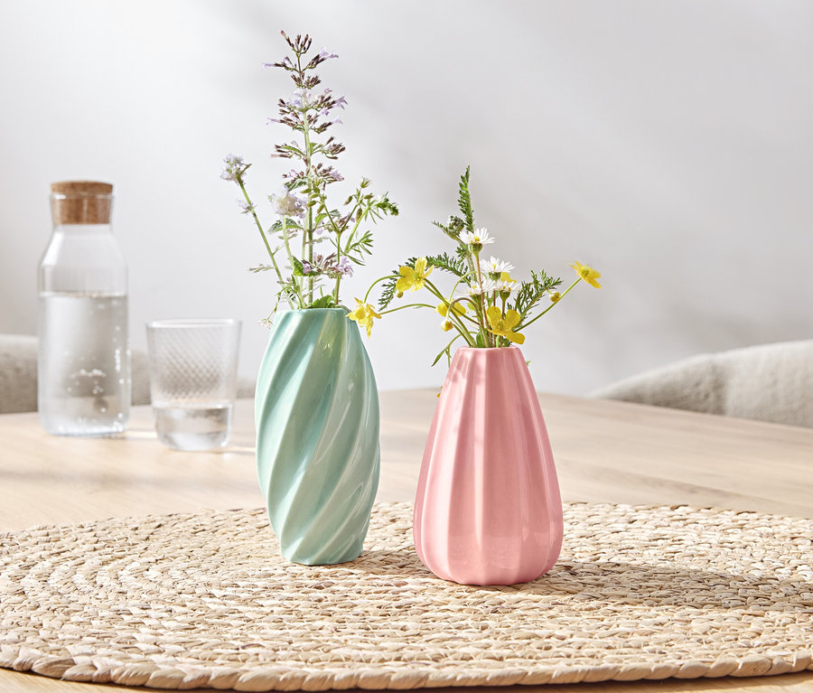 Auf einem Tisch stehen zwei Vasen mit Blumen, eine grüne und eine rosa.