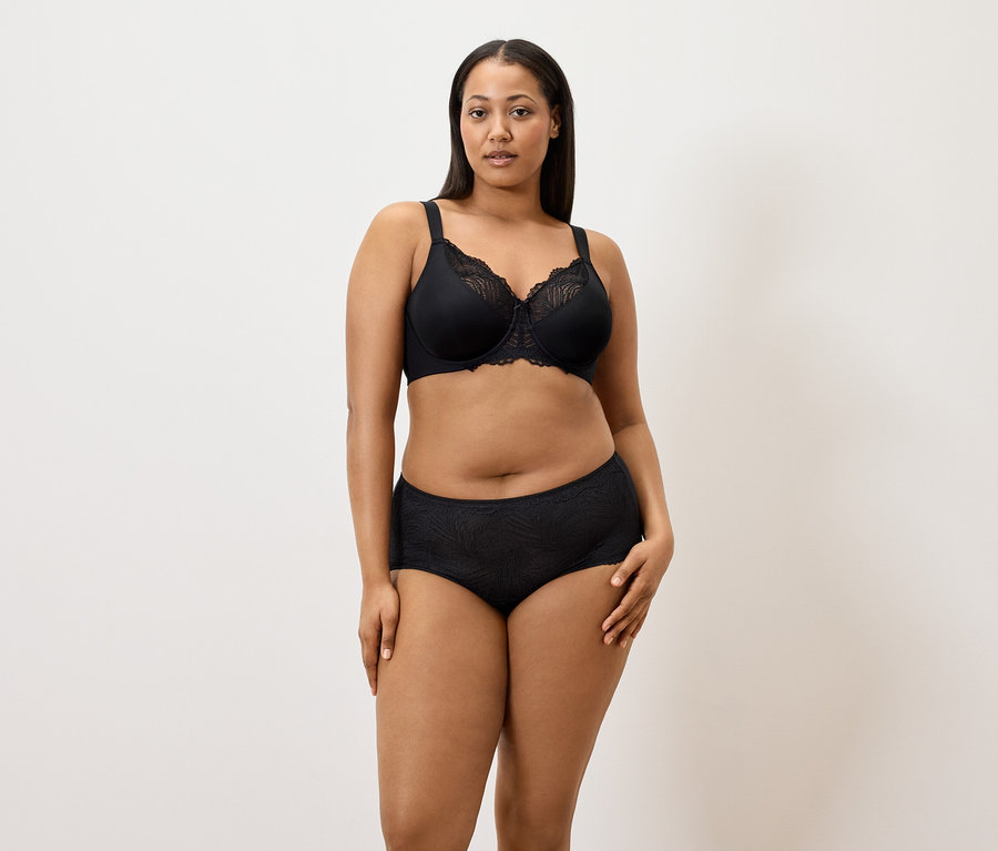 Frau trägt einen schwarzen Minimizer-BH und schwarze 5 Pantys vor einem weißen Hintergrund.