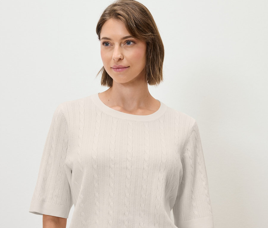 Eine Frau trägt einen cremefarbenen Zopfstrickpullover mit kurzen Ärmeln.