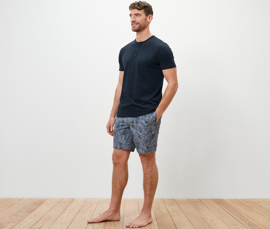 Mann im Shorty-Pyjama-Set mit gewebter Hose steht barfuß auf Holzboden.