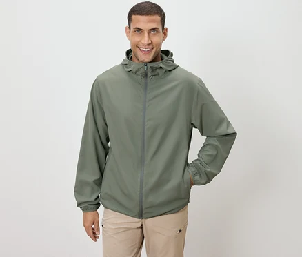 Mann mit olivgrüner Jacke und beiger Hose, eine Hand in der Tasche, vor einem weißen Hintergrund.