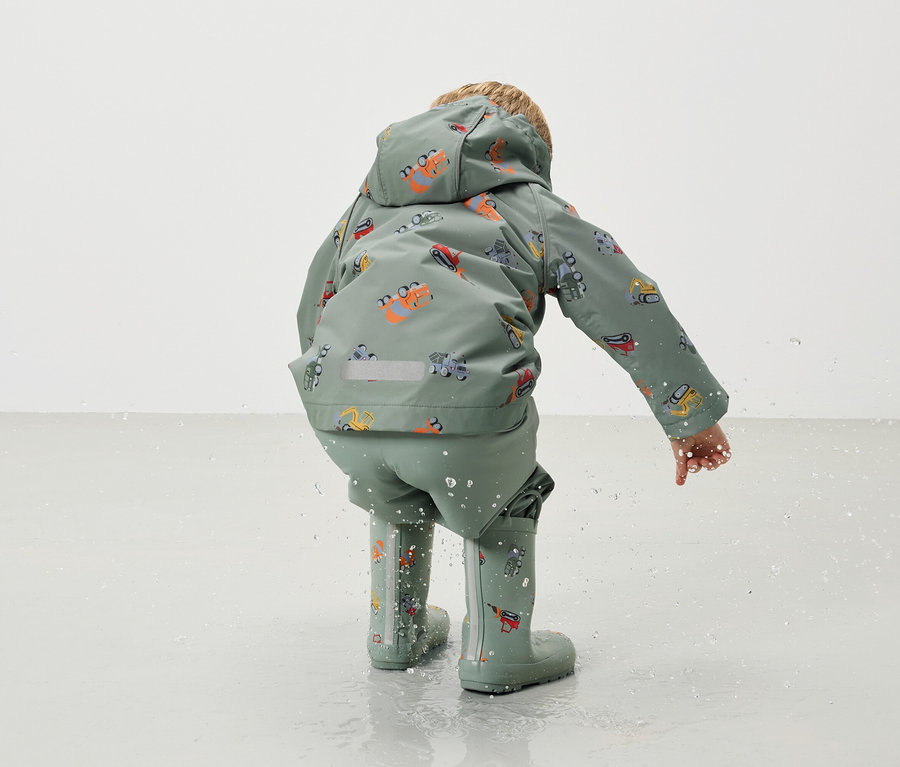 Ein Kind von hinten, trägt eine grüne Regenjacke und Regenhose mit Bagger-Motiv und passende Gummistiefel, während es im Wasser spielt.