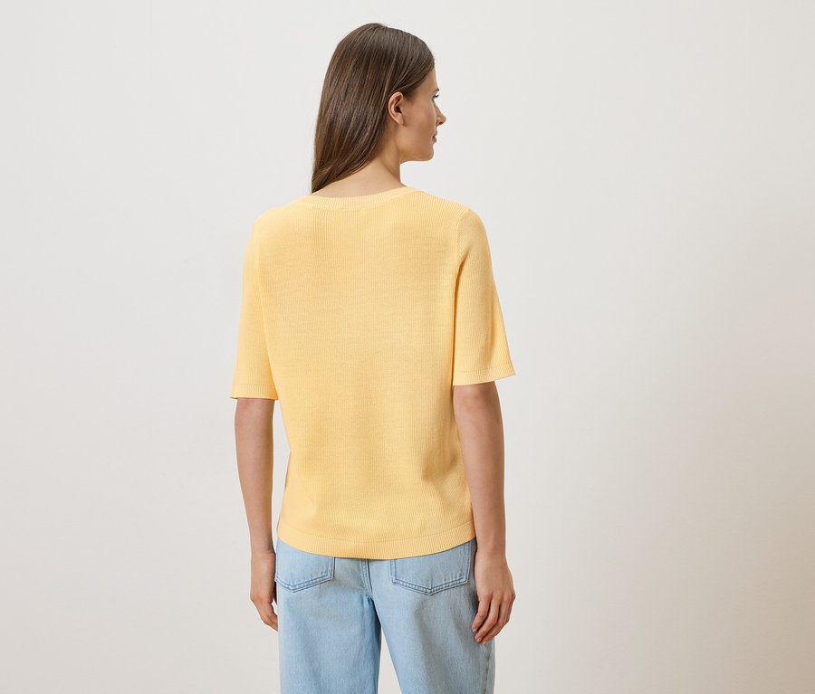 Eine Frau von hinten in hellblauen Jeans und einem gelben T-Shirt mit kurzen Ärmeln vor einem weißen Hintergrund.