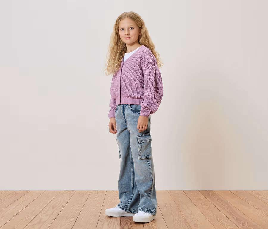 Mädchen mit blonden, lockigen Haaren, trägt eine lila Strickjacke, eine Jeanshose und weiße Schuhe.