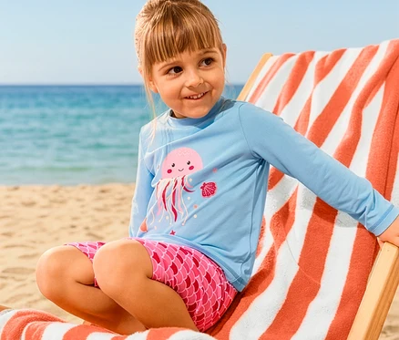 Ein Mädchen sitzt auf einem Liegestuhl mit XL-Badetuch und trägt eine Kinder-Kombi mit UV-Schutzfaktor 50+, Qualle.