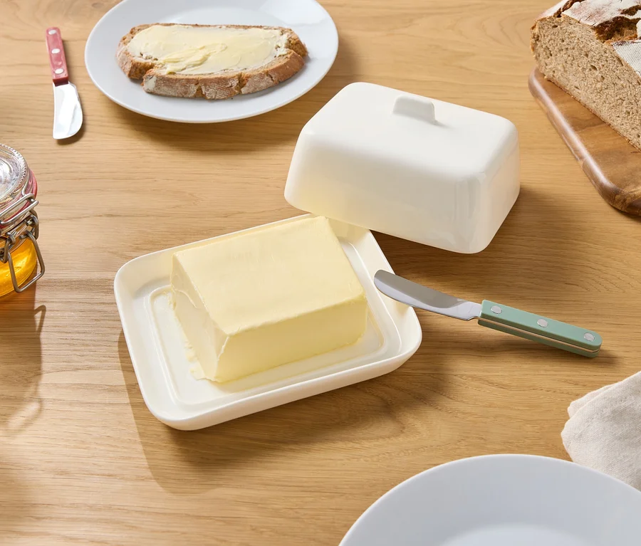 Auf einem Holztisch stehen eine geöffnete Butterdose mit Butter, ein Honigglas, ein Messer, Brot und ein Teller mit einem Butterbrot.