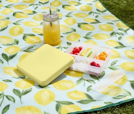 Gelbe Brotdose mit Behälter mit Obst und Gemüse und Getränk auf einer Picknickdecke mit Zitronenmuster.