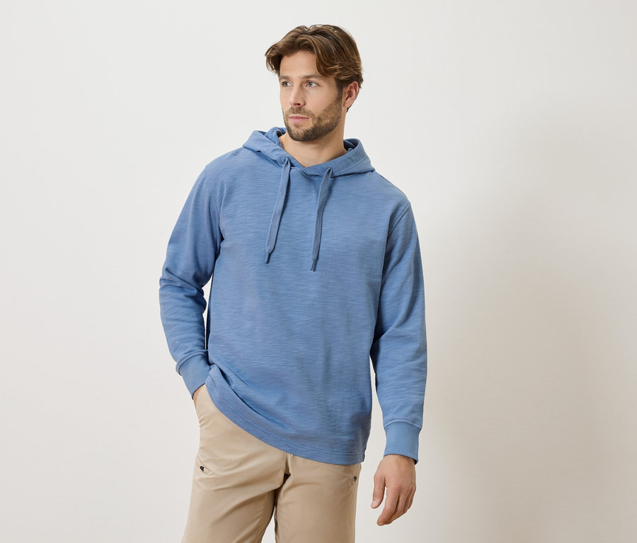 Mann mit Bart und blauem Hoodie mit Kordelzug und beigen Hosen, die Hände in den Taschen.