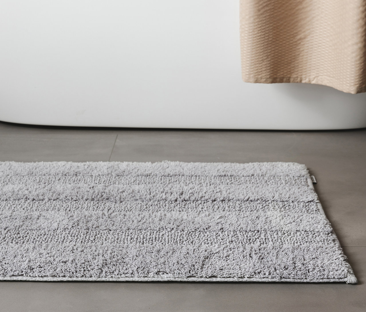 Eine hellgraue benuta Badematte »Ilka«, 50 x 80 cm, liegt vor einer weißen Badewanne auf einem grauen Boden.