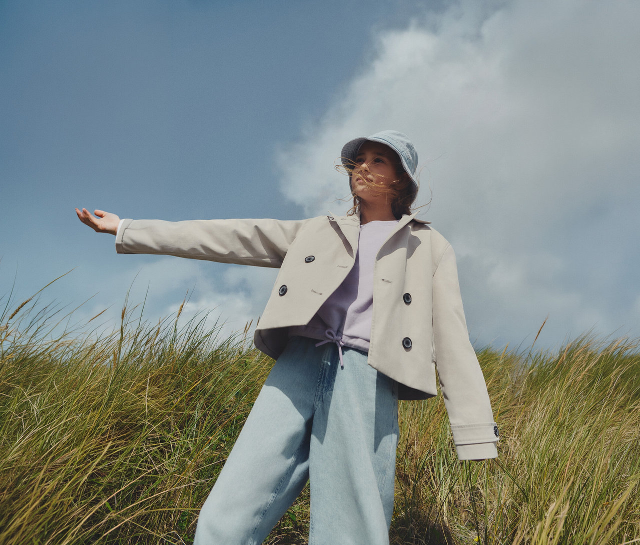 Junge Person mit Hut, beiger Jacke und Tom Tailor Kinder-Wide-Leg-Fit-Jeans mit Umschlag im Gras. Ein Arm ist ausgestreckt.