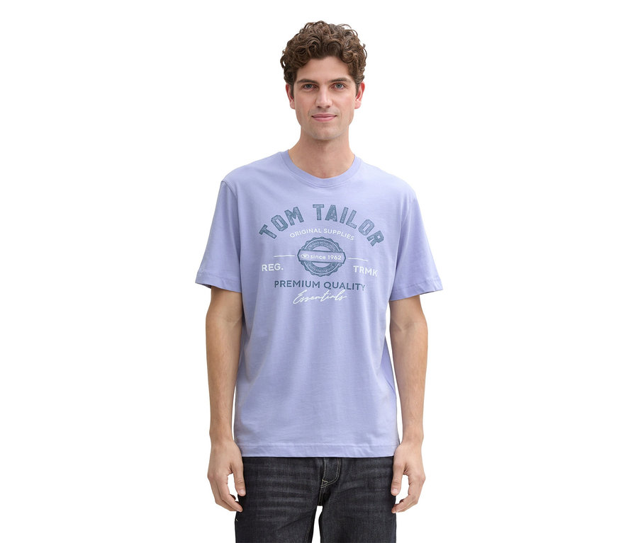 Mann mit lockigem Haar trägt ein hellblaues T-Shirt mit Tom Tailor-Logo und dunkle Jeans.