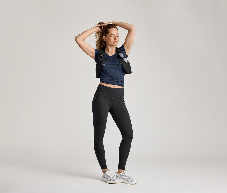 Frau posiert in Outdoor-Leggings, marineblauem Shirt und Weste vor hellem Hintergrund.