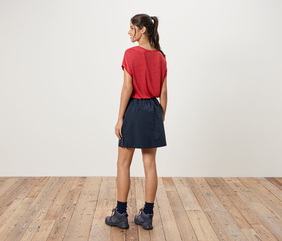 Eine Frau steht mit dem Rücken zur Kamera, sie trägt ein rotes T-Shirt, einen blauen Rock und graue Wanderschuhe.