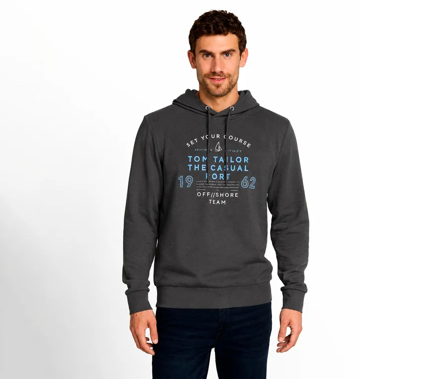Mann trägt einen dunkelgrauen Hoodie mit blau-weißem „TOM TAILOR THE CASUAL“ Grafikdruck auf der Vorderseite und dunkelblauen Jeans vor weißem Hintergrund.