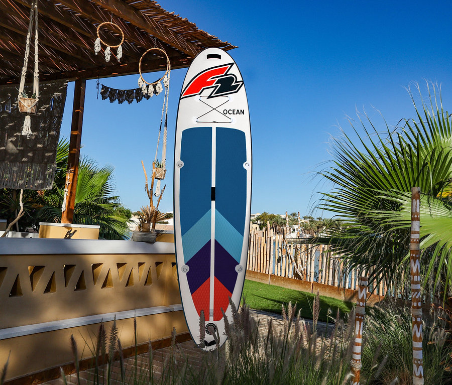 Weißes F2 SUP Board »Ocean ltd« mit blauen, hellblauen, lila und roten Mustern steht aufrecht vor einem Strohdach und Palmen unter blauem Himmel.