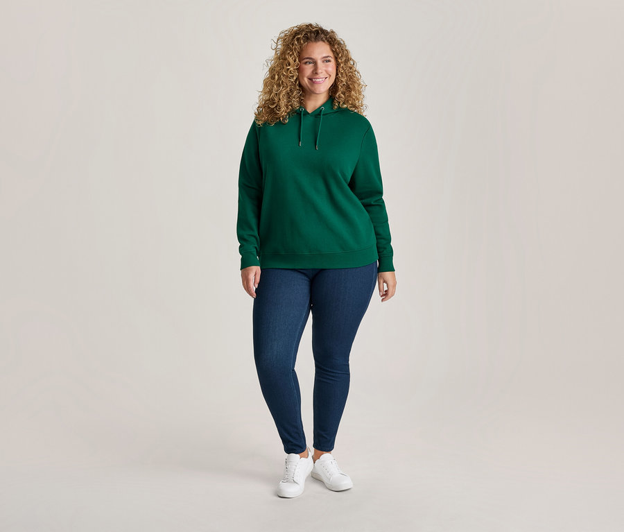 Lächelnde Frau mit lockigem Haar trägt einen grünen Hoodie, blaue Jeans und weiße Turnschuhe vor hellem Hintergrund.