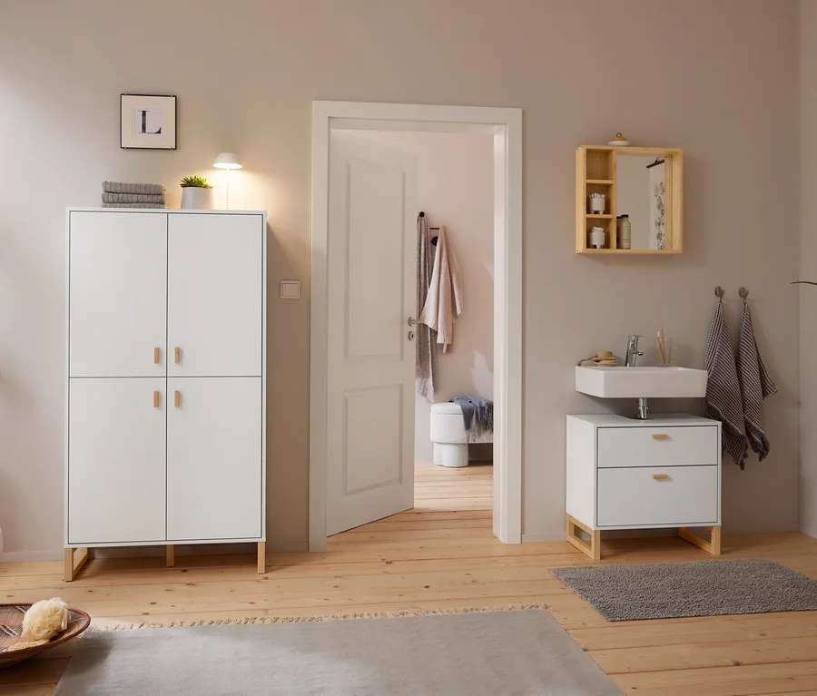 Helles Badezimmer mit Holzboden und hellgrauen Wänden. Links steht ein weißer Schrank mit einer weißen LED-Akku-Tischleuchte und einer Pflanze. Rechts hängt ein Spiegel aus massivem Bambus mit Ablagen über einem Waschbecken mit Unterschrank. Eine offene Tür führt in einen Nebenraum.