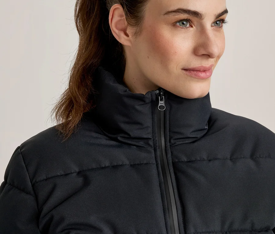 Nahaufnahme einer Frau mit zurückgebundenen Haaren, die eine schwarze Steppjacke mit hohem Kragen und Reißverschluss trägt und nach rechts blickt.