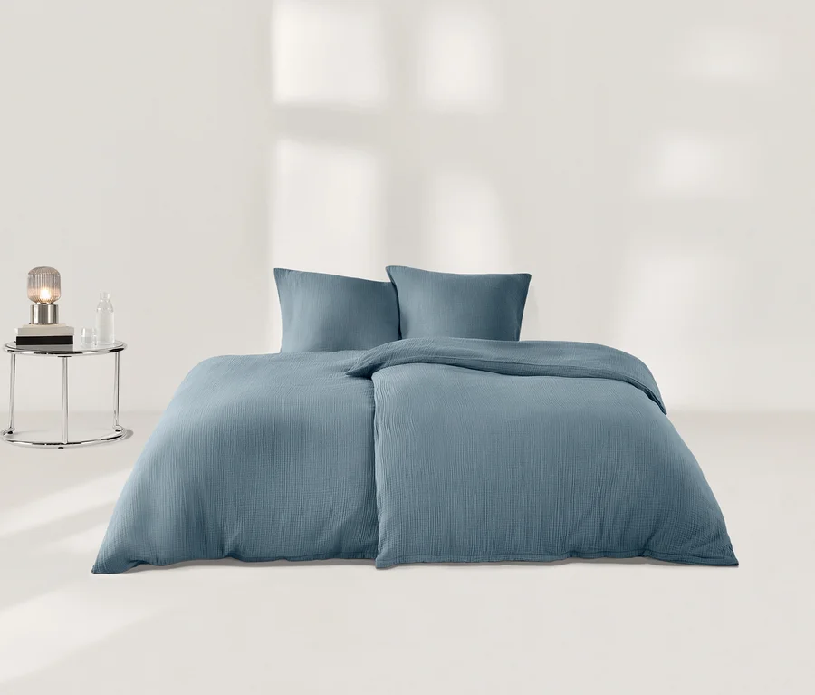 Ein Bett mit blauer Musselin-Bettwäsche und zwei Kissen. Links daneben steht ein runder Nachttisch mit einer Lampe, Büchern und einer Flasche.