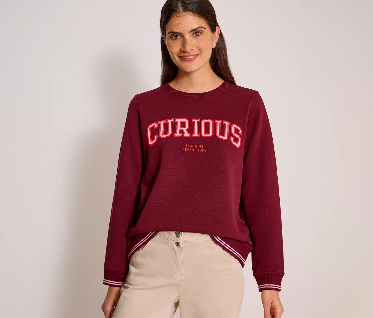 Eine lächelnde Frau mit dunklen Haaren trägt ein bordeauxrotes CECIL Sweatshirt mit der Aufschrift „CURIOUS“ in rot-weißen Buchstaben und „LEARNING NEVER STOPS“ darunter, kombiniert mit hellbeiger Hose.