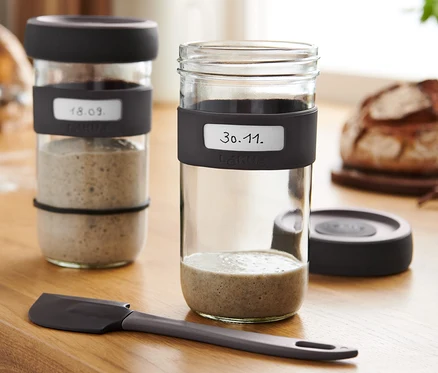 Zwei Gläser mit Sauerteig-Starter-Set auf einem Holztisch mit Brot und Spatel.