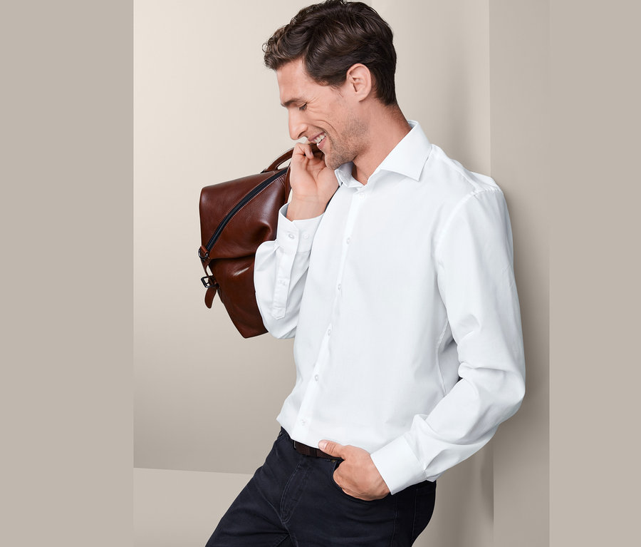 Mann lächelt und hat eine braune Tasche über die Schulter gehängt. Er trägt ein weißes Hemd, Comfort Fit, und eine schwarze Hose.